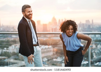 Two businesspeople standing against London rooftop view at sunset, laughing. - Powered by Shutterstock