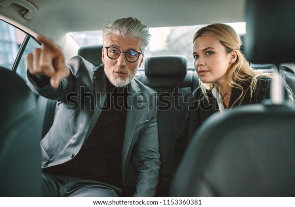 Two businesspeople traveling by car with man\
pointing away. Senior businessman showing something interesting to\
female while traveling by a\
taxi.