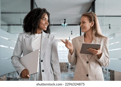 Two businesswoman working together, while walking in office and holding digital gadget pc. Small creative diversity team of African American and blond females executives meeting discuss work project. - Powered by Shutterstock