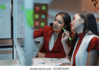 Two businesswomen brainstorming and discussing using transparent board and colorful sticky notes in a modern office at night - Powered by Shutterstock