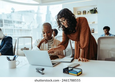 Two businesswomen collaborating in a modern office, using a laptop and having a discussion about a project. They are happy and smiling, showing their teamwork in a technology-driven workplace. - Powered by Shutterstock