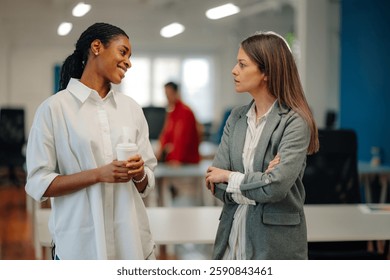 Two businesswomen are discussing work related matters during their coffee break in a modern, open plan office, fostering collaboration and communication within the team