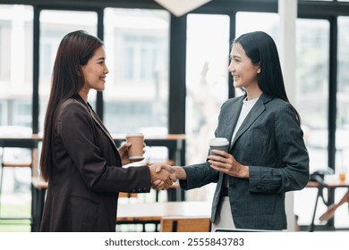 Two businesswomen in formal attire shaking hands, holding coffee cups, in a bright, modern office environment. - Powered by Shutterstock