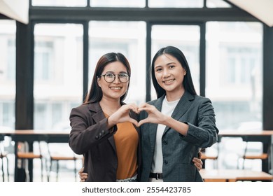 Two businesswomen in formal attire smiling and forming a heart shape with their hands in a bright office environment. - Powered by Shutterstock