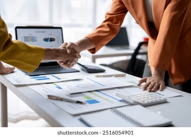 Two businesswomen are shaking hands over a desk covered with charts, graphs, and a laptop, symbolizing a successful business agreement - Powered by Shutterstock