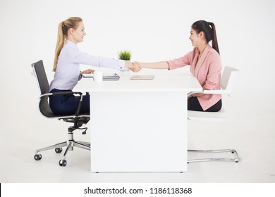 Two Businesswomen In Suits Sitting Between The Table Handshaking Of Deal On White Background. Human Resources Manager Female Interviewing Woman .interviewer Girl Shaking Hand Of During A Job Interview