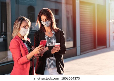 Two Businesswomen Wearing Mask And Using Phone Outside The Office