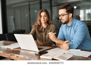 Two busy colleagues working together talking using laptop in office. Middle aged Latin female manager teaching young male worker looking at computer discussing business plan at office meeting. - Powered by Shutterstock