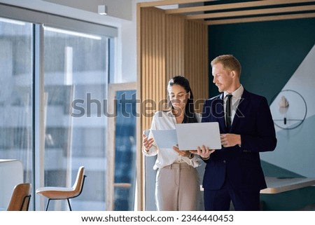 Two busy diverse coworkers discussing project walking in office with laptop and documents. International business team people colleagues standing having teamwork conversation. Authentic shot
