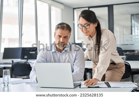 Two busy diverse professional coworkers discussing work using laptop in office. Asian employee learning online project discussing business plan with mature manager looking at computer at meeting.