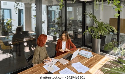 Two busy happy business women of young and middle age talking in green office sitting at desk. Professional ladies employee and manager having conversation using laptop at work. Candid top view. - Powered by Shutterstock