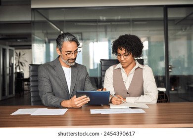 Two busy happy diverse professional business team people working in office using digital tablet. Older male executive manager talking to young female colleague having conversation sitting at work desk - Powered by Shutterstock