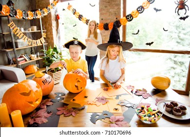 Two busy kids in halloween headwear are making hand made decor for party, mum is watching them from behind, relaxing with hot drink, cup of coffee, desk top with yellow candles, sweets, fall leaves