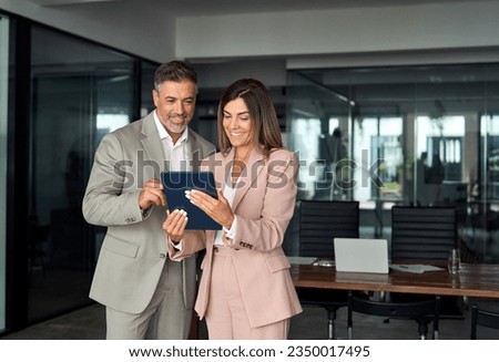 Two busy older professional corporate business executives man and woman wearing suits holding tablet technology device having discussion working on digital project plan standing in office at meeting.