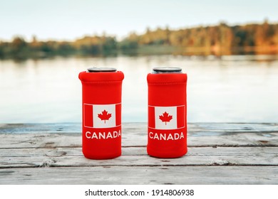 Two Cans Of Beer In Red Cozy Beer Can Cooler With Canadian Flag Standing On Wooden Pier By Lake Outdoor. Friends Celebrating Canada Day National Celebration On July 1 In Nature Park By Water. 