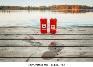 Two Cans Of Beer In Red Cozy Beer Can Coolers With Canadian Flag Standing On Wooden Pier By Lake Outdoors. Wet Footprints On Wooden Dock. Friends Celebrating Canada Day National Celebration By Water. 