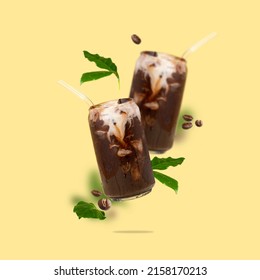 Two can-shaped glass with iced coffee, coffee beans and leaves in the air on a yellow background. Cold coffee with non-dairy milk. Modern and creative composition of flying summer vegeterian drink.