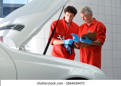 Two car mechanics wearing red overalls and protective gloves standing in the garage and looking at car engine while talking to each other - Powered by Shutterstock