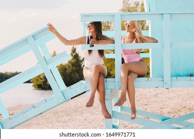 Two Carefree Women Friends Drinking Cold Lemonade And Laughing While Sitting On Lifeguard Tower. Girl Pointing Away, Calling Someone For Party. Happy Girls Cheers With Coke Soft Drinks On Sunny Beach