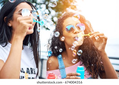 Two Carefree Young Girlfriends Having Fun Together Blowing Bubbles With A Toy Bubble Wand While Enjoying A Sunny Day Together Outside 