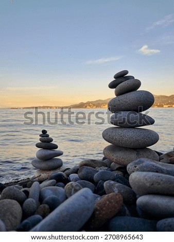 Two carefully balanced stacks of smooth, grey stones (cairns) stand on a pebble beach by the calm sea, symbolizing balance, meditation, or tranquility.