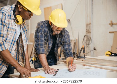 Two carpenter man wear helmet meeting planning job together at carpentry workshop, craftsmen talking checklist and plan on blueprint paper in woodshop, teamwork, National Carpenters Day