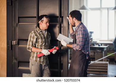 Two carpenters are talking while holding papers and ruler. One of them is wearing safety mask. Carpentry shop background - Shutterstock ID 520703374
