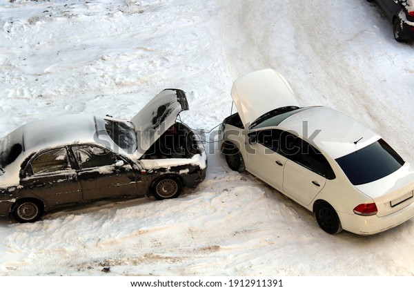 two cars stand in the winter with open hoods. One\
car charges another