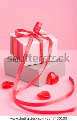 Two carton gift boxes of silver and beige colour with red bow made of textile ribbon laying on pastel pink background with heart shaped decorations prepared for celebration of valentines day. Vertical