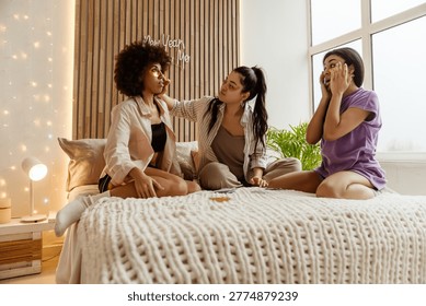 Two casually dressed Black, and one Asian women in their late 20s sit next to each other on a bed, in a decorated with string lights sunlit bedroom, applying under-eye patches near a big window.