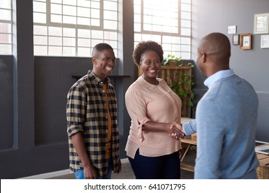 Two casually dressed young African business colleagues standing in a large modern office smiling and shaking hands together while another coworker looks on