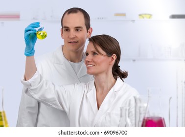 Two Caucasian Biotechnicians In The Lab, Examining Yellow Liquid In A Conical Flask