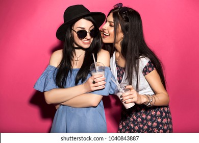 Two caucasian brunette hipster woman in casual stylish outfit having fun drinking milkshakes with straws. They standing on a bright pink background. They gossip with a cheerful, happy emotions - Powered by Shutterstock