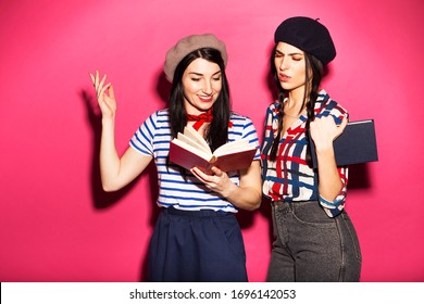 Two Caucasian Brunette Hipster Woman In Casual Stylish French Outfit With Beret, Having Fun Reading And Peaking In Each Other Books. They Standing On A Bright Pink Background. Cheerful, Happy Emotions