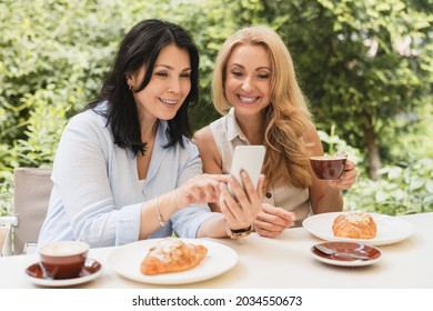 Two caucasian businesswomen sisters best friends girlfriends showing news social media photos on smart phone cellphone sitting in cafe and drinking coffee during breakfast. - Powered by Shutterstock