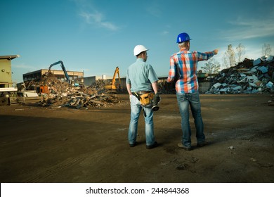 two caucasian engineers standing in recycling center outdoors, pointing at pile of scrap metal - Powered by Shutterstock