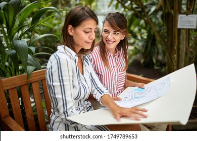 Two Caucasian Female Landscape Architects Sitting On Wooden Bench In Botanical  Garden, Watching Draft Design For Arranging Botanic Park