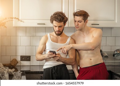 two caucasian gays use smartphone, stand together in kitchen. lovely married homosexual couple look at screen of mobile phone and discuss message from their mutual friend - Powered by Shutterstock