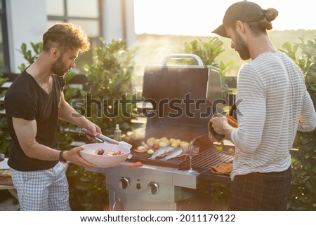Two caucasian guys cooking vegetables and fish on a modern gas grill at backyard on a sunset. Healthy eating on the open air, male friendship