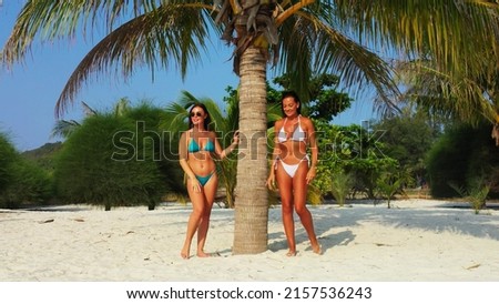 A two Caucasian pretty females in bikini standing next to a coconut palm tree on a sandy beach, Koh Samui, Thailand