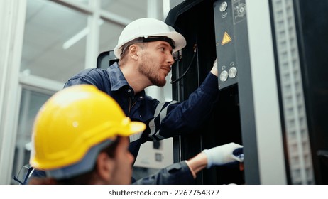 Two Caucasian production engineers in safety wear are assisting in adjusting and maintaining CNC machine in the factory. Male factory workers are examining the industrial machine to find an error.