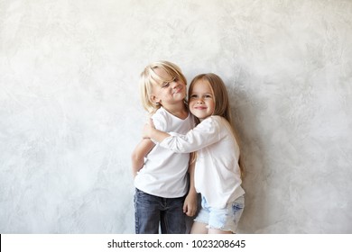 Two Caucasian Siblings Brother And Sister Posing For Picture During Family Photo Shooting: Stubborn Blonde Boy Doesn't Want To Embrace His Little Sister, Grimacing And Hiding Arms Behind His Back