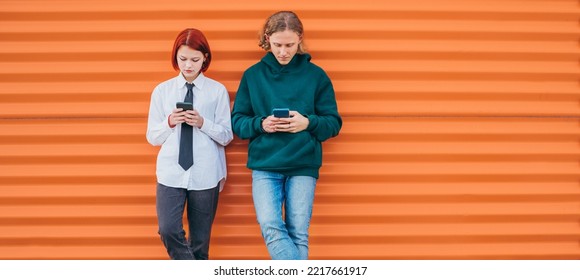 Two Caucasian Teen Friends Boy And A Girl Browsing Their Smartphone Devices While They Lean On The Orange Wall Background. Careless Young Teenhood 
 Time And A Modern Technology Concept Image.