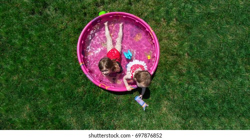 Two Caucasian Toddler Brown Haired Girls Plays In A Pink Backyard Kiddie Pool During  A Hot Summer Day