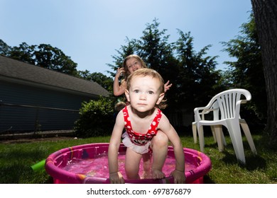 Two Caucasian Toddler Brown Haired Girls Plays In A Pink Backyard Kiddie Pool During  A Hot Summer Day