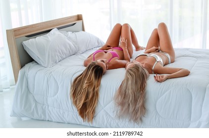 Two Caucasian unrecognizable unknown beautiful blonde hair hot female fashion models in pink and silver sexy lingerie underwear laying down on white sheet blanket bed with pillows in bedroom together.