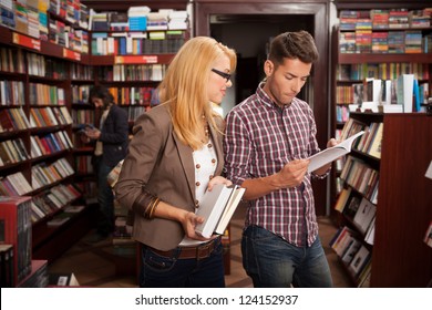 Two Caucasian Young People In A Bookstore Reading Something In A Book With Many Bookshelves In The Background