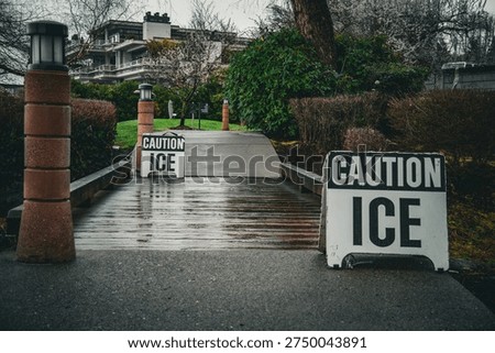 Two caution ice signs are placed on a wet wooden walkway leading towards a building, warning of slippery conditions