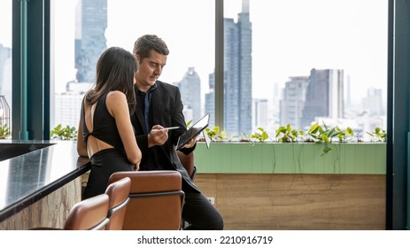 Two CEOs Have A Conversation Over A Coffee Bar With Cityscape Background. A Smart Couple Stand Next To A Bar Counter In An Open Space. Male And Female Managers In Black Dress For Formal Dinner.