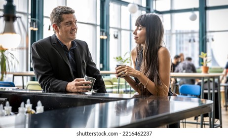 Two CEOs Have A Conversation Over A Coffee Bar With Cityscape Background. A Smart Couple Stand Next To A Bar Counter In An Open Space. Male And Female Managers In Black Dress For Formal Dinner.